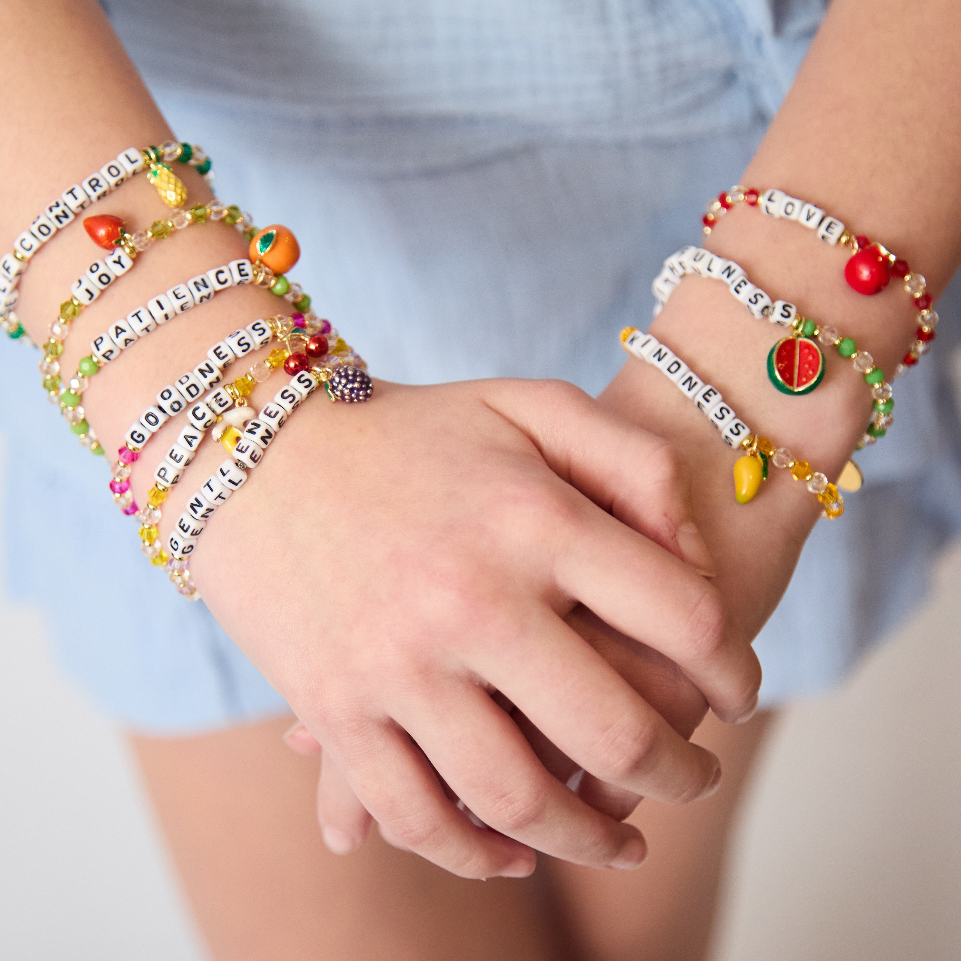 Close-up of hands wearing colorful bracelets with various charms and text like Love, Gentleness, Patience, Joy, Self Control, Goodness, and Kindness.